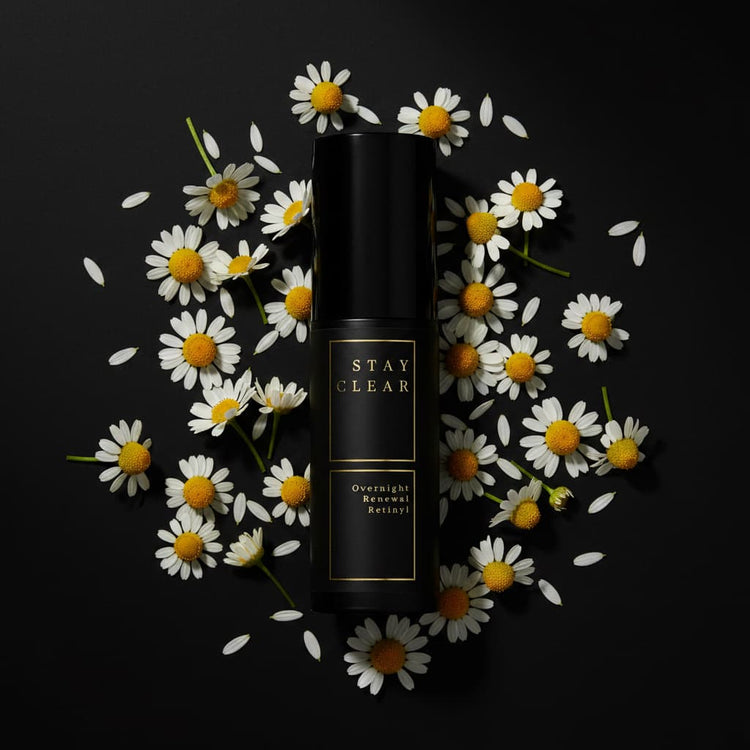 Stay Clear Beauty Overnight Renewal Retinyl bottle surrounded by fresh chamomile flowers on a dark background — retinaldehyde night treatment for skin renewal, fine lines, uneven texture, and acne-prone skin.
