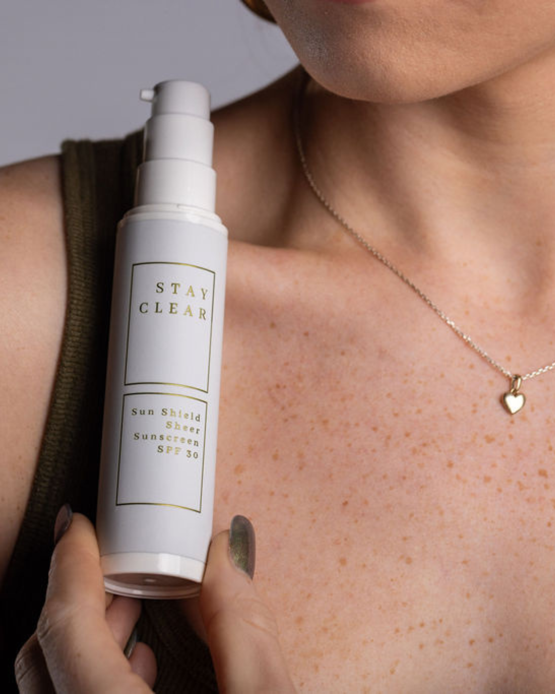 Women wearing a gold heart necklace, beautiful lightly freckled skin, holding a white bottle Sun Shield Sheer Sunscreen SPF 30 - Stay Clear Beauty