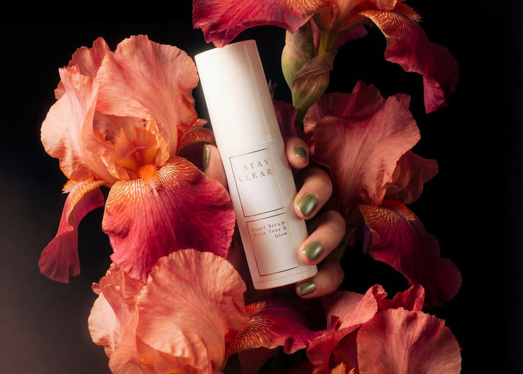Hand with green nail polish holding Stay Clear Beauty Super Serum Even Tone & Glow Sensitive surrounded by pink and peach flowers — brightening serum for dark spots, post-acne marks, and uneven skin tone on sensitive skin.
